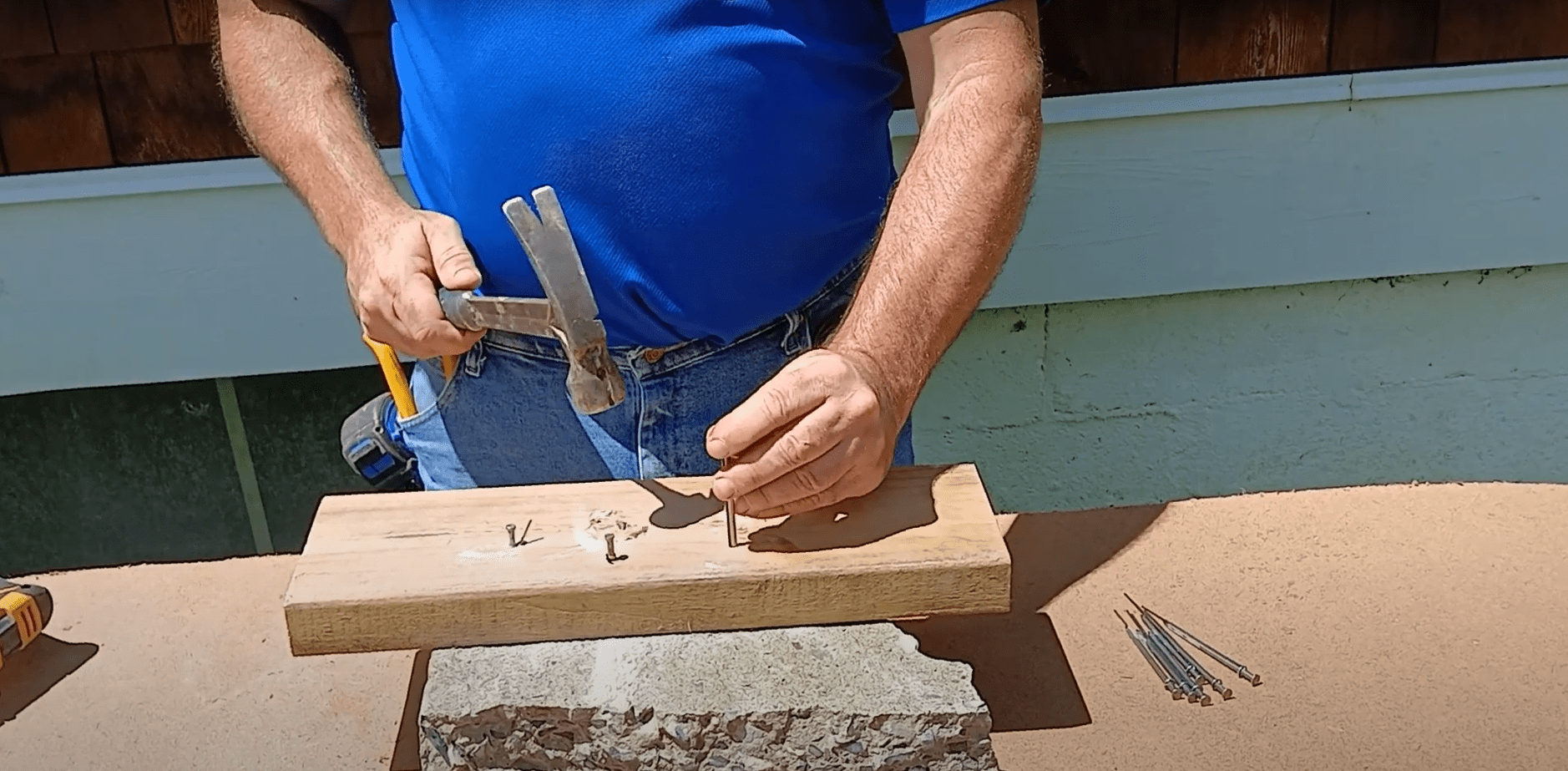 A person in a blue shirt hammers a nail into a wooden plank on a work surface, surrounded by tools and additional nails, demonstrating a DIY project.